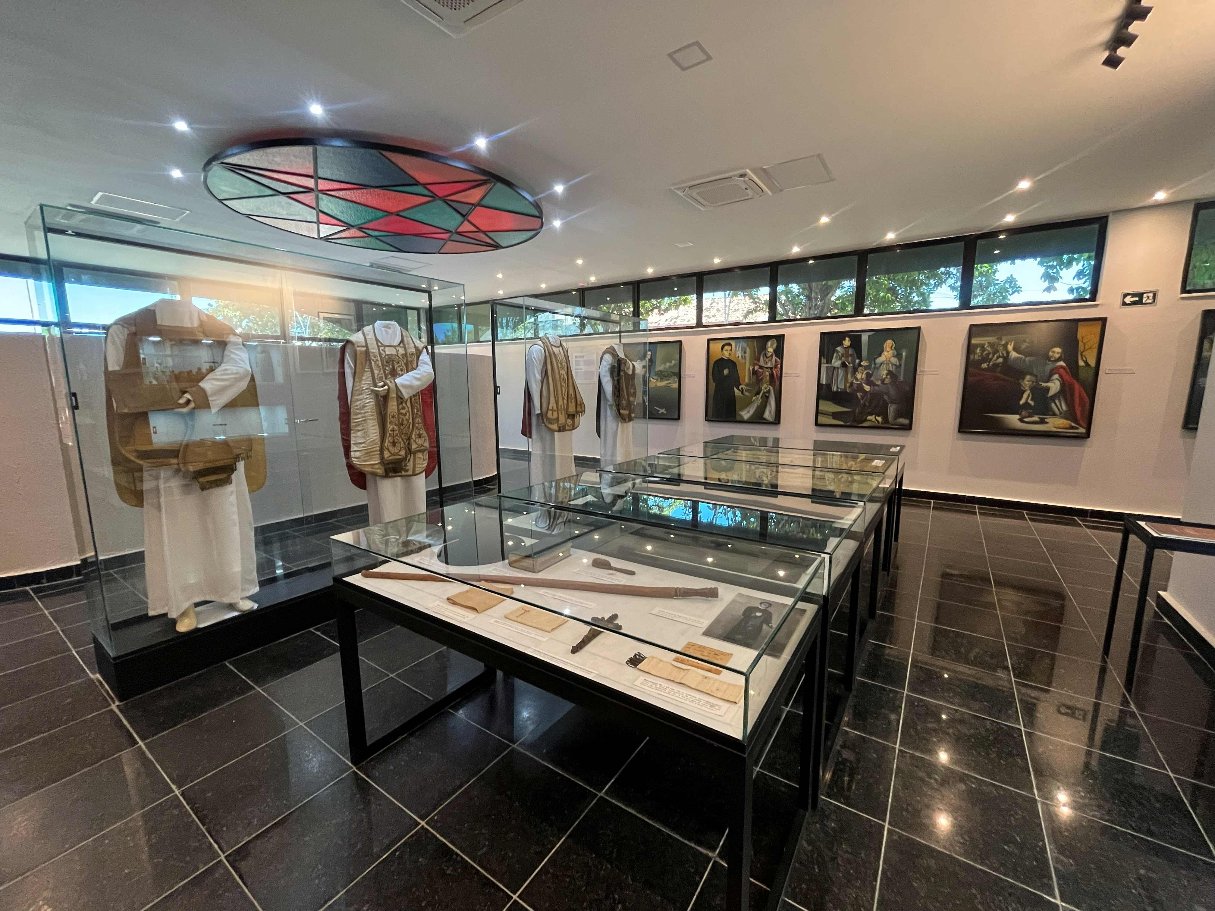 Sala de exposição do Memorial Padre Cícero com vestimentas litúrgicas, objetos históricos em vitrines de vidro e pinturas religiosas nas paredes.